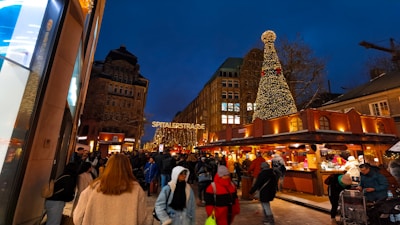 A busy street scene at night, featuring a brightly lit Christmas market stall and a large Christmas tree adorned with lights. A crowd of people is walking and shopping along the street, with historic buildings in the background. Festive decorations and holiday lights create a warm, welcoming atmosphere.