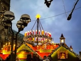 A festive Christmas market stall adorned with a large Advent wreath featuring red candles and garlands. The ambience is enhanced by decorative lights and a wooden, gingerbread-style structure bearing the name Käthe Wohlfahrt. Several people are gathered around, enjoying the holiday atmosphere under the dim evening sky.