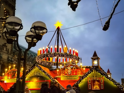 A festive Christmas market stall adorned with a large Advent wreath featuring red candles and garlands. The ambience is enhanced by decorative lights and a wooden, gingerbread-style structure bearing the name Käthe Wohlfahrt. Several people are gathered around, enjoying the holiday atmosphere under the dim evening sky.