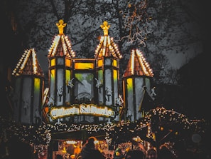 A festive building with illuminated turrets and warm yellow lights, set against a dark evening sky. The structure has decorative elements, including small figures and intricate designs. Below, a crowd gathers beneath the lights, indicating a lively atmosphere.