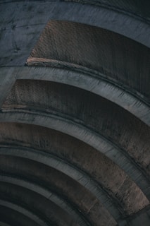 Several large, curved concrete beams or arches form an abstract pattern. The structure appears to be industrial, with signs of weathering and wear. The lighting emphasizes the texture and shadows of the material.