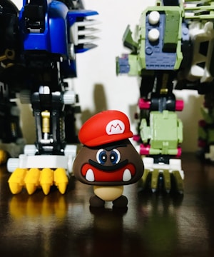 A small figurine with a red hat and a cartoonish face stands in front of two large robot-like toys. The robots have intricate details with colors like blue, green, and yellow, and appear to be made of plastic or metal parts.