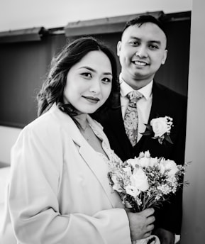 A black and white photograph of a couple standing closely together. The woman is holding a bouquet of flowers and is wearing a coat over a dress, while the man is dressed in a suit with a floral tie and a rose boutonniere. Both are smiling softly and looking toward the camera, conveying a sense of warmth and connection.