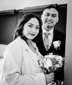 A black and white photograph of a couple standing closely together. The woman is holding a bouquet of flowers and is wearing a coat over a dress, while the man is dressed in a suit with a floral tie and a rose boutonniere. Both are smiling softly and looking toward the camera, conveying a sense of warmth and connection.
