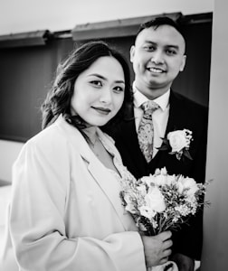 A black and white photograph of a couple standing closely together. The woman is holding a bouquet of flowers and is wearing a coat over a dress, while the man is dressed in a suit with a floral tie and a rose boutonniere. Both are smiling softly and looking toward the camera, conveying a sense of warmth and connection.