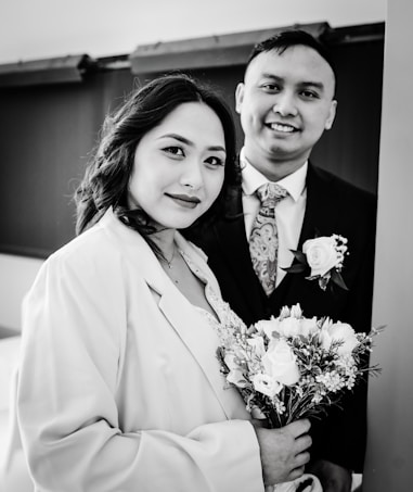 A black and white photograph of a couple standing closely together. The woman is holding a bouquet of flowers and is wearing a coat over a dress, while the man is dressed in a suit with a floral tie and a rose boutonniere. Both are smiling softly and looking toward the camera, conveying a sense of warmth and connection.