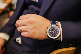 A hand wearing a luxury wristwatch and a silver ring. The person is dressed in a dark suit with a white shirt cuff visible. The wristwatch has a blue leather strap and a detailed blue dial with multiple indicators. The setting appears to be indoors.