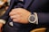 A hand wearing a luxury wristwatch and a silver ring. The person is dressed in a dark suit with a white shirt cuff visible. The wristwatch has a blue leather strap and a detailed blue dial with multiple indicators. The setting appears to be indoors.