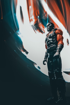 A dramatic image featuring a muscular action figure standing against a vibrant, abstract background with swirling red, blue, and white colors. The figure is posed powerfully, clad in a dark outfit with red elements on the arms.