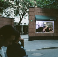 A brick wall with a fashion advertisement displayed, featuring a person lying down in a black outfit. The background includes trees and a building, with two people walking in the foreground.