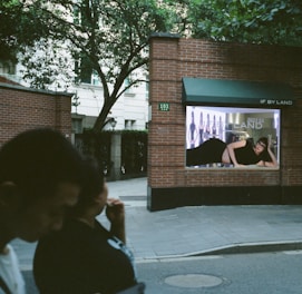 A brick wall with a fashion advertisement displayed, featuring a person lying down in a black outfit. The background includes trees and a building, with two people walking in the foreground.