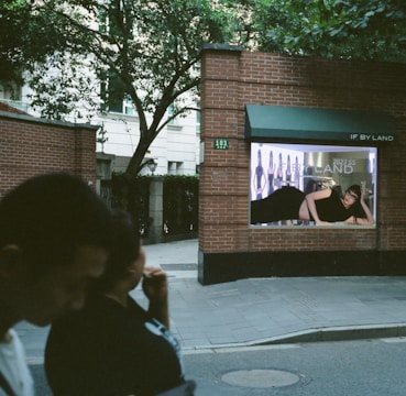 A brick wall with a fashion advertisement displayed, featuring a person lying down in a black outfit. The background includes trees and a building, with two people walking in the foreground.