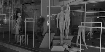 A display window showcasing multiple featureless mannequins arranged in standing and leaning positions. Some mannequins are positioned on platforms, while others are laying or missing body parts. The setting appears to be an empty store with metal racks and minimal decoration, creating an industrial and stark atmosphere.