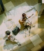 A pair of musicians are performing indoors. One person is playing an acoustic guitar and singing into a microphone while seated. Another musician is playing a hand drum or cajon. The floor is a polished surface with a pattern of light and shadow.