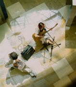A pair of musicians are performing indoors. One person is playing an acoustic guitar and singing into a microphone while seated. Another musician is playing a hand drum or cajon. The floor is a polished surface with a pattern of light and shadow.