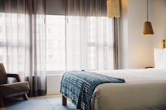 A modern bedroom with soft natural light coming through sheer curtains. The bed is neatly made with a white duvet and a blue patterned throw. There's a brown armchair near the window and a hanging lamp above a bedside table. The room has a calm and minimalist design.