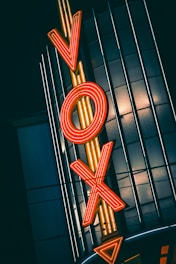 A bold, retro-style neon sign with the letters 'VOX' is illuminated in bright orange and set against a dark, vertical-striped building facade.