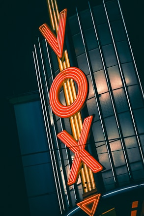 A bold, retro-style neon sign with the letters 'VOX' is illuminated in bright orange and set against a dark, vertical-striped building facade.