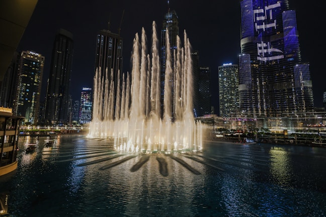 A night shot of Dubai Fountain with illuminated water jets and crowds.