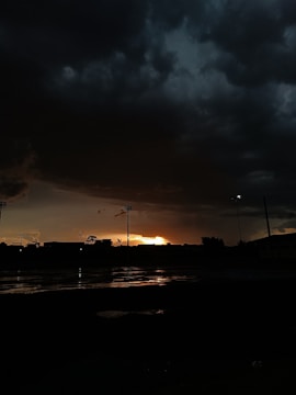 A dramatic scene with dark, heavy clouds filling the sky, creating a looming atmosphere. The horizon reveals a sliver of golden sunlight breaking through, casting reflections on a wet surface below. Silhouettes of structures and trees are visible against the dimly lit background.