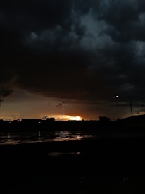 A dramatic scene with dark, heavy clouds filling the sky, creating a looming atmosphere. The horizon reveals a sliver of golden sunlight breaking through, casting reflections on a wet surface below. Silhouettes of structures and trees are visible against the dimly lit background.