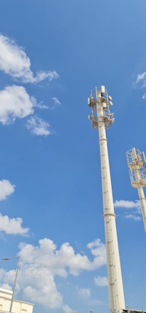 Two tall communication towers rise against a bright blue sky scattered with fluffy white clouds. The towers are equipped with multiple antennas and equipment, indicating their use for telecommunications. At the bottom left, part of a modern building and a streetlight are visible.