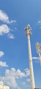 Two tall communication towers rise against a bright blue sky scattered with fluffy white clouds. The towers are equipped with multiple antennas and equipment, indicating their use for telecommunications. At the bottom left, part of a modern building and a streetlight are visible.