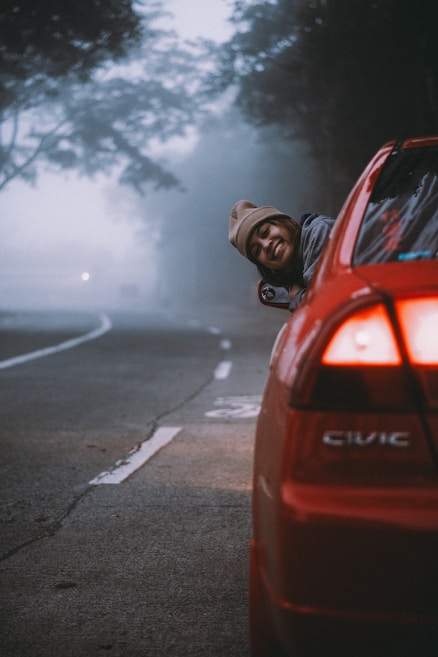 A person wearing a beanie leans out of the window of a red car on a foggy road, smiling. The road is lined with trees shrouded in mist.