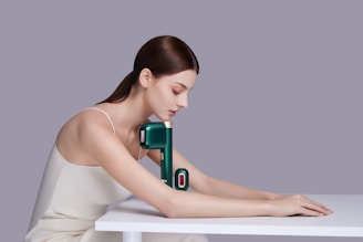 A woman with long brown hair wearing a white sleeveless top is posed resting her arms and head over a white table. Positioned against her arm is a sleek, modern massage device in a dark green color with a gold accent.