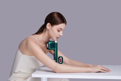 A woman with long brown hair wearing a white sleeveless top is posed resting her arms and head over a white table. Positioned against her arm is a sleek, modern massage device in a dark green color with a gold accent.
