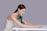 A woman with long brown hair wearing a white sleeveless top is posed resting her arms and head over a white table. Positioned against her arm is a sleek, modern massage device in a dark green color with a gold accent.
