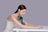 A woman with long brown hair wearing a white sleeveless top is posed resting her arms and head over a white table. Positioned against her arm is a sleek, modern massage device in a dark green color with a gold accent.