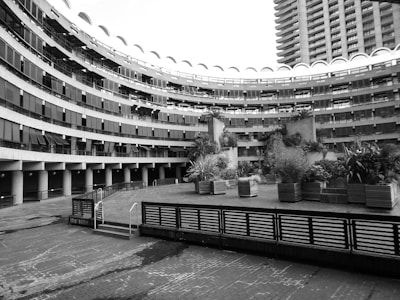 A curved, multi-story building features numerous balconies and a series of arch-shaped windows along the top. In the foreground, there is a landscaped garden area with a variety of plants in rectangular planters. The architecture displays a modern, industrial style with concrete and metal elements.