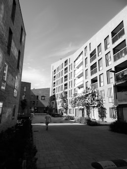 A modern urban courtyard is surrounded by multi-story residential buildings with balconies and large windows. A solitary person walks down the paved pathway. Trees and shrubs are present, adding a touch of greenery to the architectural setting.