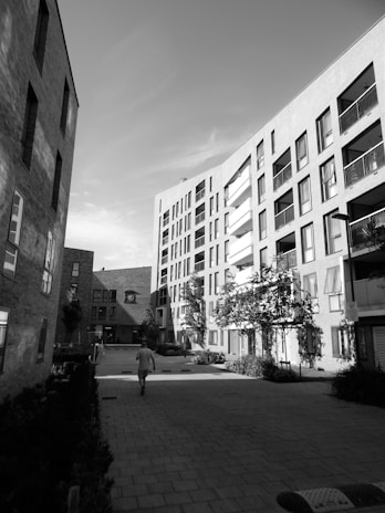 A modern urban courtyard is surrounded by multi-story residential buildings with balconies and large windows. A solitary person walks down the paved pathway. Trees and shrubs are present, adding a touch of greenery to the architectural setting.
