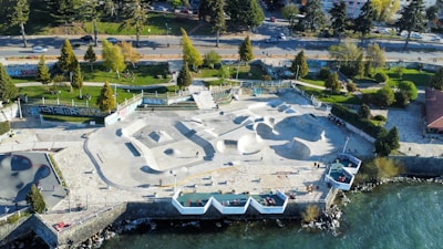 Aerial view of an outdoor skate park located by a waterfront. The park features multiple skate ramps and bowls, surrounded by trees and greenery. Alongside the skatepark, there is a walkway with scattered benches. People are seen engaging in skateboarding activities. The surrounding area includes a road with moving vehicles and a building to the right. Graffiti is visible on some walls.