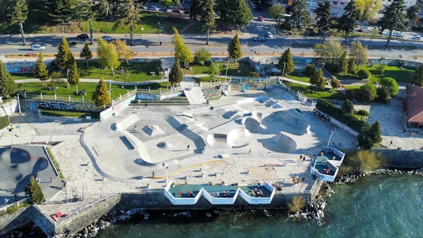 Aerial view of an outdoor skate park located by a waterfront. The park features multiple skate ramps and bowls, surrounded by trees and greenery. Alongside the skatepark, there is a walkway with scattered benches. People are seen engaging in skateboarding activities. The surrounding area includes a road with moving vehicles and a building to the right. Graffiti is visible on some walls.