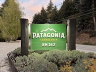 A vibrant green sign displays the words 'Patagonia Cerveceria KM 24.7' with a mountain graphic above the text. The sign is set against a backdrop of lush, green trees and vegetation, bordered by wooden poles, with a gravel road visible beside it.
