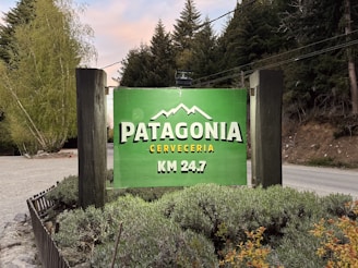 A vibrant green sign displays the words 'Patagonia Cerveceria KM 24.7' with a mountain graphic above the text. The sign is set against a backdrop of lush, green trees and vegetation, bordered by wooden poles, with a gravel road visible beside it.