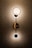 An elegant wall light fixture with a modern design is mounted on a patterned wallpaper. The lamp consists of two circular lights connected by a vertical rod, casting a warm glow that highlights the intricate texture of the background.
