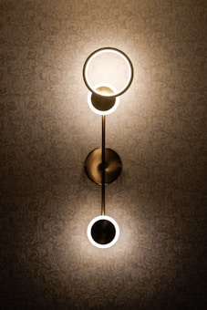 An elegant wall light fixture with a modern design is mounted on a patterned wallpaper. The lamp consists of two circular lights connected by a vertical rod, casting a warm glow that highlights the intricate texture of the background.