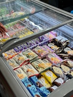 An assortment of packaged ice cream and frozen desserts is displayed inside a commercial freezer with a glass lid. Various brands and flavors are arranged neatly in rows, featuring colorful packaging with labels in different languages. A hand is visible near the top left corner, possibly reaching towards one of the items.