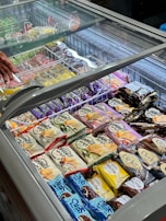 An assortment of packaged ice cream and frozen desserts is displayed inside a commercial freezer with a glass lid. Various brands and flavors are arranged neatly in rows, featuring colorful packaging with labels in different languages. A hand is visible near the top left corner, possibly reaching towards one of the items.