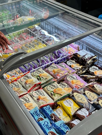 An assortment of packaged ice cream and frozen desserts is displayed inside a commercial freezer with a glass lid. Various brands and flavors are arranged neatly in rows, featuring colorful packaging with labels in different languages. A hand is visible near the top left corner, possibly reaching towards one of the items.