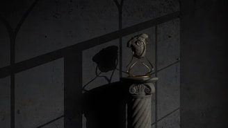 A textured wall serves as the backdrop for a stone pedestal holding an anatomically detailed heart sculpture made of stone or similar material. The heart is enclosed in a delicate, gold-colored metal frame. Shadows of a window grille are cast on the wall, creating a dramatic play of light and dark.