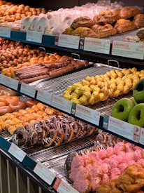 A display of various pastries and donuts is arranged on metal shelves. There are multiple types of pastries including croissants, twisty breadsticks, and a selection of colorful donuts with chocolate and pink icing topped with sprinkles. Labels with prices and descriptions in a foreign language are visible beneath the items.