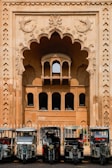 In front of an intricately designed historical building with an ornate archway featuring carved fish motifs, several auto rickshaws are parked in a row. The building showcases traditional architectural elements with arched windows and detailed stonework. The scene suggests a blend of cultural heritage and modern transportation.