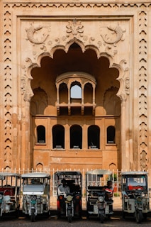 In front of an intricately designed historical building with an ornate archway featuring carved fish motifs, several auto rickshaws are parked in a row. The building showcases traditional architectural elements with arched windows and detailed stonework. The scene suggests a blend of cultural heritage and modern transportation.