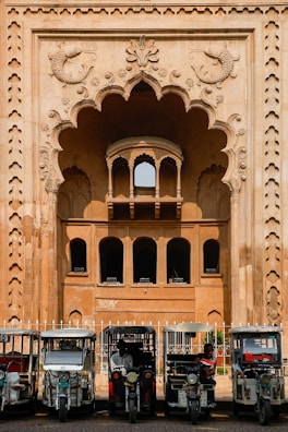 In front of an intricately designed historical building with an ornate archway featuring carved fish motifs, several auto rickshaws are parked in a row. The building showcases traditional architectural elements with arched windows and detailed stonework. The scene suggests a blend of cultural heritage and modern transportation.
