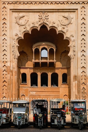 In front of an intricately designed historical building with an ornate archway featuring carved fish motifs, several auto rickshaws are parked in a row. The building showcases traditional architectural elements with arched windows and detailed stonework. The scene suggests a blend of cultural heritage and modern transportation.